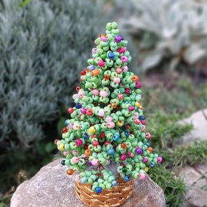 May include: A handmade beaded Christmas tree with green beads forming the foliage and colorful beads as ornaments. The tree sits in a gold-colored basket on a stone. The background is blurred with green foliage.
