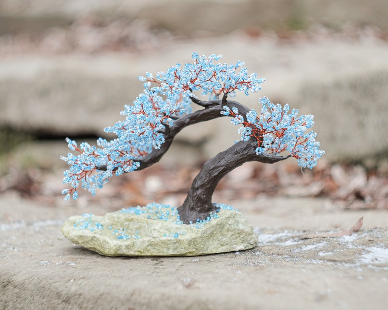 May include: A blue beaded bonsai tree with a brown wooden base, sitting on a light gray stone surface.