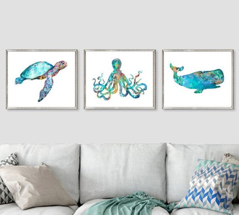 May include: Three framed watercolor prints featuring sea creatures: a sea turtle, an octopus, and a whale. The artwork uses shades of blue, green, and yellow against a white background. The prints are displayed above a white couch.