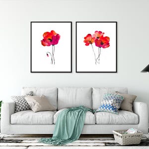 May include: Two framed watercolour paintings of red poppies on a white background. The paintings are hanging on a white wall above a white sofa with cushions and a throw blanket.