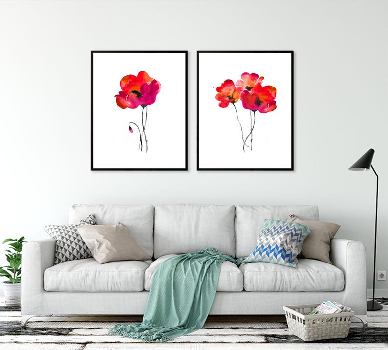 May include: Two framed watercolour paintings of red poppies on a white background. The paintings are hanging on a white wall above a white sofa with cushions and a throw blanket.