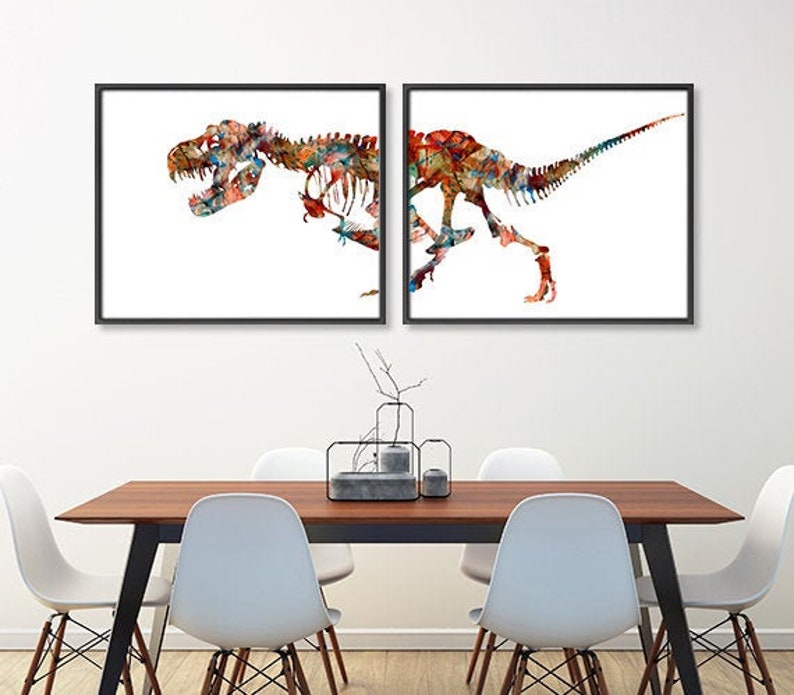 Może przedstawiać: Dwa oprawione wydruki artystyczne przedstawiające kolorowe ilustracje szkielet&oacute;w dinozaur&oacute;w. Grafika przedstawia T-Rexa w żywym stylu akwarelowym, z mieszanką czerwonych, niebieskich i ż&oacute;łtych odcieni. Wydruki są prezentowane na białej ścianie nad drewnianym stołem.