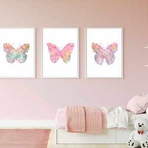 May include: Three watercolor butterfly prints in pink, blue, and purple tones. The butterflies are on a white background and are framed in white frames. The prints are hanging on a pink wall.