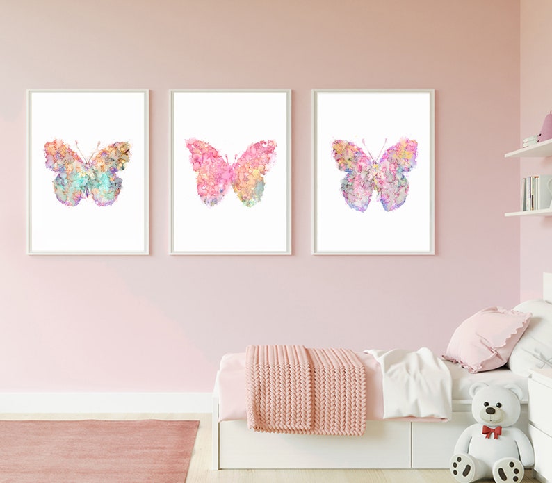 May include: Three watercolor butterfly prints in pink, blue, and purple tones. The butterflies are on a white background and are framed in white frames. The prints are hanging on a pink wall.