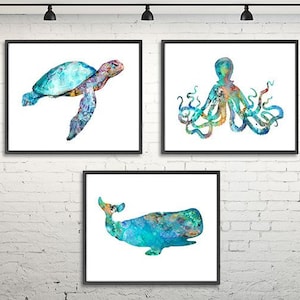 May include: Three framed watercolor paintings of sea creatures: a sea turtle, an octopus, and a whale. The artwork features shades of blue, green, and orange against a white background. The frames are black, and the art is displayed on a white brick wall.