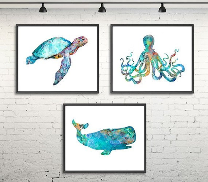 May include: Three framed watercolor paintings of sea creatures: a sea turtle, an octopus, and a whale. The artwork features shades of blue, green, and orange against a white background. The frames are black, and the art is displayed on a white brick wall.