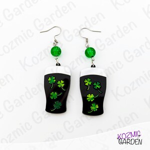 May include: A pair of St. Patrick's Day themed earrings. Each earring features a black pint glass design with white foam and green shamrocks. The earrings are accented with a green bead and silver-tone hooks.