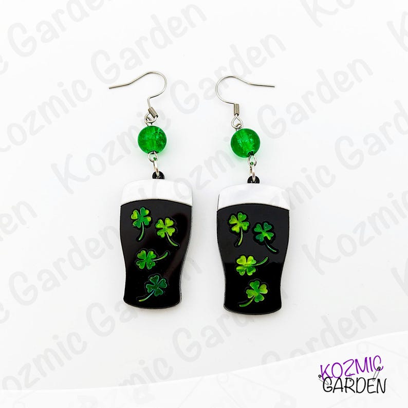 May include: A pair of St. Patrick's Day themed earrings. Each earring features a black pint glass design with white foam and green shamrocks. The earrings are accented with a green bead and silver-tone hooks.