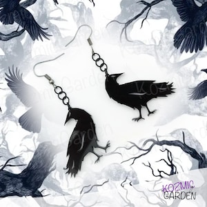 Puede incluir: Un par de pendientes de cuervo negro con cadenas plateadas. Los cuervos están en silueta sobre un fondo blanco con un diseño de rama de árbol en blanco y negro.