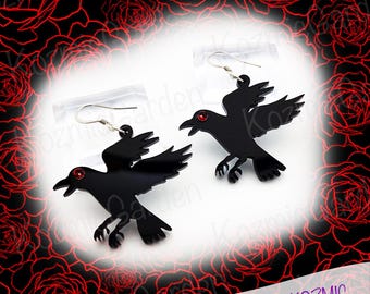 May include: A pair of black crow earrings with red eyes. The earrings are made of acrylic and have a dangling design.