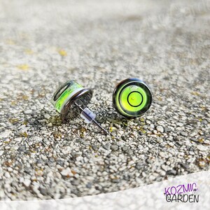 A pair of silver stud earrings with a green and black spirit level bubble in the centre. The earrings are on a grey textured surface.