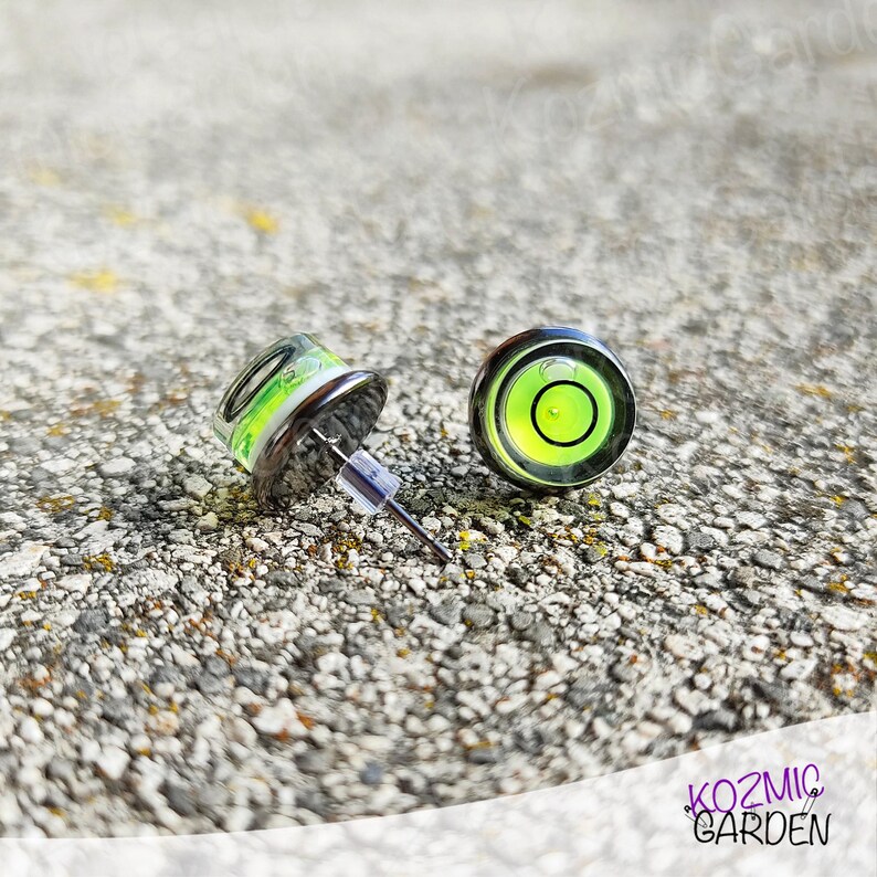 A pair of silver stud earrings with a green and black spirit level bubble in the centre. The earrings are on a grey textured surface.