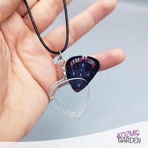 May include: A black guitar pick necklace with a Guns N' Roses design. The pick is attached to a clear heart-shaped pendant and a black cord necklace. The pendant is held by a hand against a light blue background.
