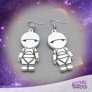 May include: A pair of silver earrings featuring a white robot design with green eyes. The robots are stylized with a simple, rounded head and a body with lines.