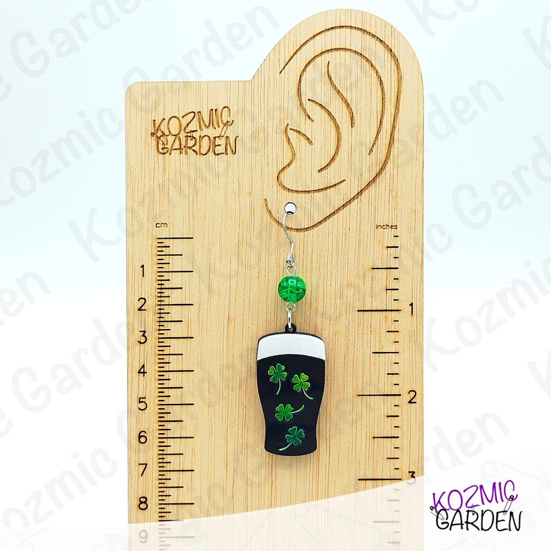 May include: A single dangle earring featuring a black pint glass design with three green shamrocks. The earring has a green bead and a silver-colored hook. The earring is displayed on a wooden measuring board with the text "KOZMIC GARDEN".