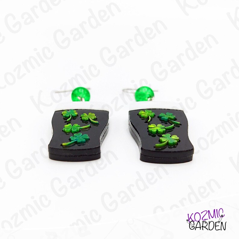 May include: A pair of black earrings shaped like a pint of stout, with green shamrocks and a white foam top. Each earring has a green bead at the top and a silver hook. The earrings are from Kozmic Garden.