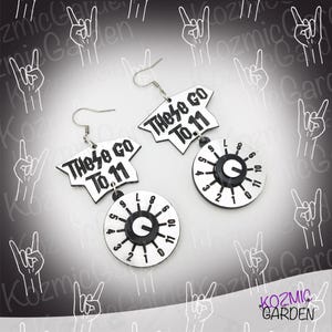 May include: A pair of novelty earrings. Each earring features a white, t-shirt-shaped top with the text "These Go To 11" in black. Below, a circular dial with numbers 1-11 and a black center. The earrings are silver-toned.