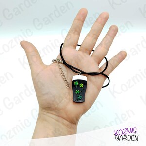 May include: A black pendant necklace featuring a pint of stout with a white head and green shamrocks. The pendant hangs from a black cord with a silver-tone chain extender. The necklace is a St. Patrick's Day themed accessory.