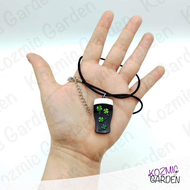 May include: A black pendant necklace featuring a pint of stout with a white head and green shamrocks. The pendant hangs from a black cord with a silver-tone chain extender. The necklace is a St. Patrick's Day themed accessory.