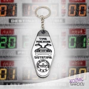 Könnte beinhalten: Ein silberfarbener Schlüsselanhänger mit einem schwarz-weißen Design. Der Schlüsselanhänger zeigt die Wörter "TIME MACHINE" und "OUTATIME" mit einer Autoillustration. Er hat auch eine silberne Kette und einen Ring.
