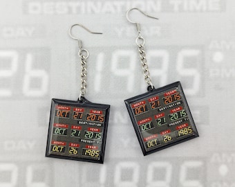 May include: A pair of Back to the Future-themed earrings. Each earring features a black square with a digital display showing the date "OCT 21 2015" and "OCT 26 1985". The earrings hang from silver-colored hooks and chains.