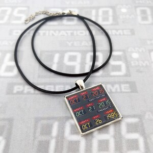 May include: A black cord necklace featuring a square pendant with a digital display graphic. The display shows three rows of dates: OCT 21 2015 under DESTINATION TIME, OCT 21 2015 under PRESENT TIME, and OCT 26 1985 under LAST TIME DEPARTED.