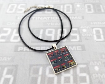 May include: A black cord necklace featuring a square pendant with a digital display graphic. The display shows three rows of dates: OCT 21 2015 under DESTINATION TIME, OCT 21 2015 under PRESENT TIME, and OCT 26 1985 under LAST TIME DEPARTED.