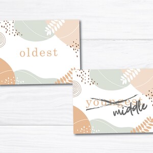 May include: Two cards with abstract designs in shades of brown, beige, and green. The text "oldest" is on one card and the text "youngest" is crossed out with the text "middle" written over it on the other card.
