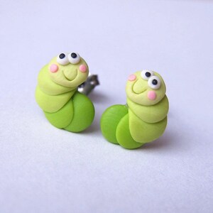 May include: A pair of green and white worm-shaped earrings with black eyes and pink cheeks. The earrings are made of polymer clay and have a silver post.