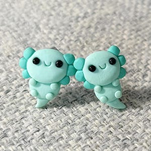 Könnte beinhalten: Ein Paar türkisblaue Axolotl-Ohrringe aus Polymer Clay. Die Axolotl haben schwarze Augen und ein kleines Lächeln.