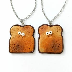 Könnte beinhalten: Zwei silberfarbene Ketten mit toastförmigen Anhängern. Jeder Anhänger ist in Braun- und Beigetönen texturiert, hat zwei weiße Wackelaugen und einen dunkelbraunen Rand. Ein Anhänger zeigt einen traurigen Mund, der andere ein lächelndes Gesicht.