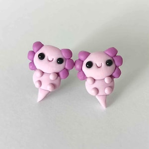 May include: Two handmade axolotl earrings in pink. Each earring has a smiling face with black eyes and a frilly, purple head. The axolotls have small arms and a pointed tail. These cute earrings are ideal for anyone who loves unique, handmade jewellery.