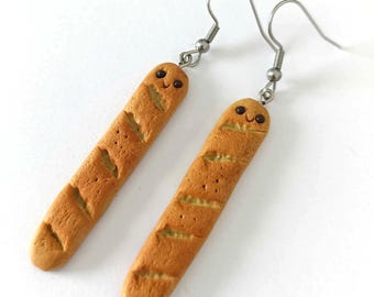 May include: A pair of novelty earrings featuring miniature baguette bread loaves with smiling faces. The earrings are a light brown color with dark brown details and silver-colored ear wires.