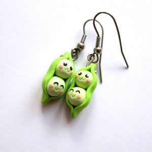 Pode incluir: Um par de brincos de vagem de ervilha verde com rostos de desenhos animados fofos. Os brincos são feitos de argila polimérica e têm ganchos prateados.