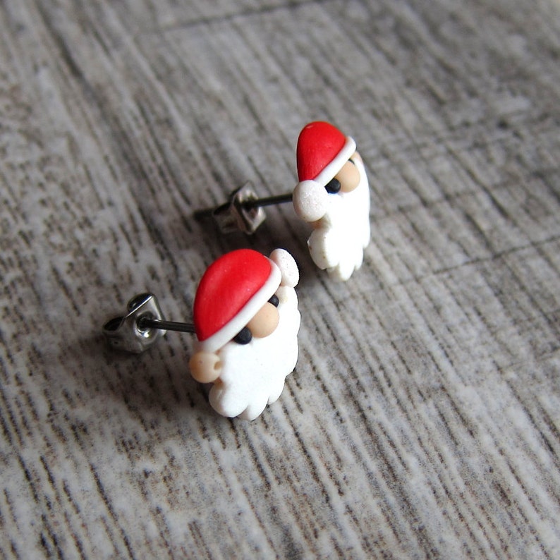 May include: A pair of silver stud earrings featuring a white Santa Claus head with a red hat. The Santa Claus head is made of polymer clay.