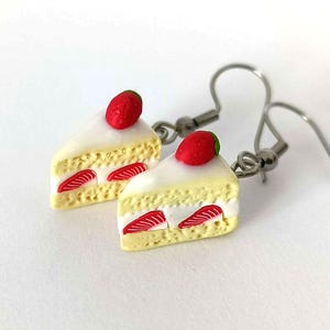 Könnte beinhalten: Ein Paar ausgefallene Ohrringe in Form von Miniatur-Erdbeerkuchenstücken. Jeder Ohrring hat einen cremefarbenen Kuchenboden, weißen Zuckerguss, rote Erdbeerscheiben und eine rote Erdbeere oben drauf. Die Ohrringe haben silberfarbene Fischhakenverschlüsse.