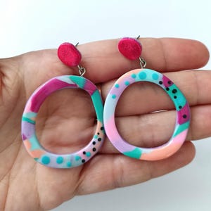 Puede incluir: Un par de pendientes colgantes con aros circulares irregulares con un patrón multicolor de rosa, verde azulado, lavanda y melocotón, acentuado por pequeños puntos negros. Cada pendiente tiene un poste circular superior de color rosa brillante.