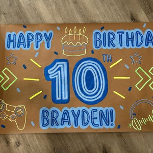 May include: A brown birthday banner with blue and white text that reads "HAPPY BIRTHDAY 10TH BRAYDEN!". The banner includes illustrations of a cake, stars, game controller, and headphones.