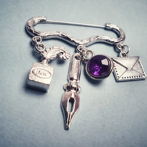May include: A silver-toned safety pin brooch featuring four dangling charms: a square inkwell with the word INK, a fountain pen nib, a round faceted purple gemstone in a silver setting, and a square envelope with a flap and dotted border detail.