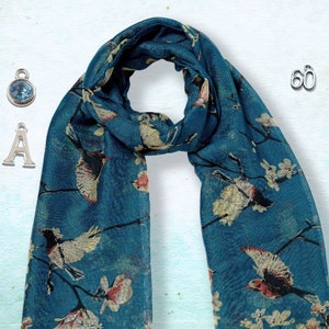 May include: A teal blue scarf with a white floral and bird print. The scarf is shown with three silver charms: a letter A, a number 60, and a blue gemstone.