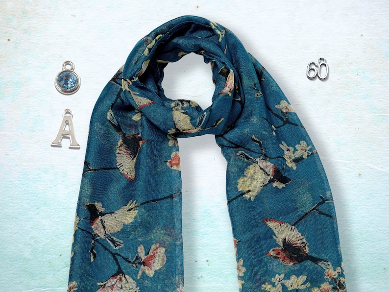May include: A teal blue scarf with a white floral and bird print. The scarf is shown with three silver charms: a letter A, a number 60, and a blue gemstone.