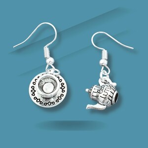 May include: Silver-toned dangle earrings. One earring features a teacup on a saucer, the other a teapot. The teapot has the words "Tea Time" etched on it. Both earrings have a decorative design and are suspended from a silver hook.