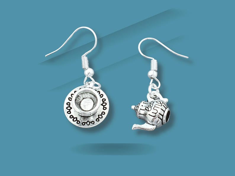 May include: Silver-toned dangle earrings. One earring features a teacup on a saucer, the other a teapot. The teapot has the words "Tea Time" etched on it. Both earrings have a decorative design and are suspended from a silver hook.