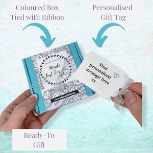May include: A turquoise gift box with a white and gray patterned design. The box is tied with a white ribbon and has a white gift tag attached. The gift tag has the text "Your personalised message here XX" and a small heart symbol.