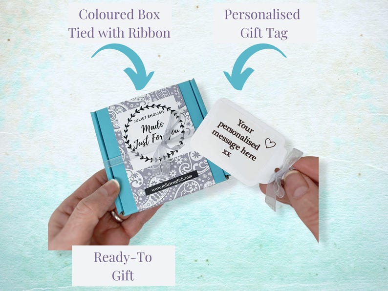 May include: A turquoise gift box with a white and gray patterned design. The box is tied with a white ribbon and has a white gift tag attached. The gift tag has the text "Your personalised message here XX" and a small heart symbol.