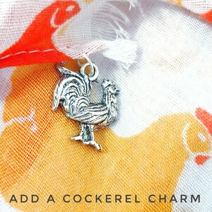 May include: A silver rooster charm with a detailed design, against a background of a white fabric with a chicken print. The text "ADD A COCKEREL CHARM" is visible at the bottom of the image.