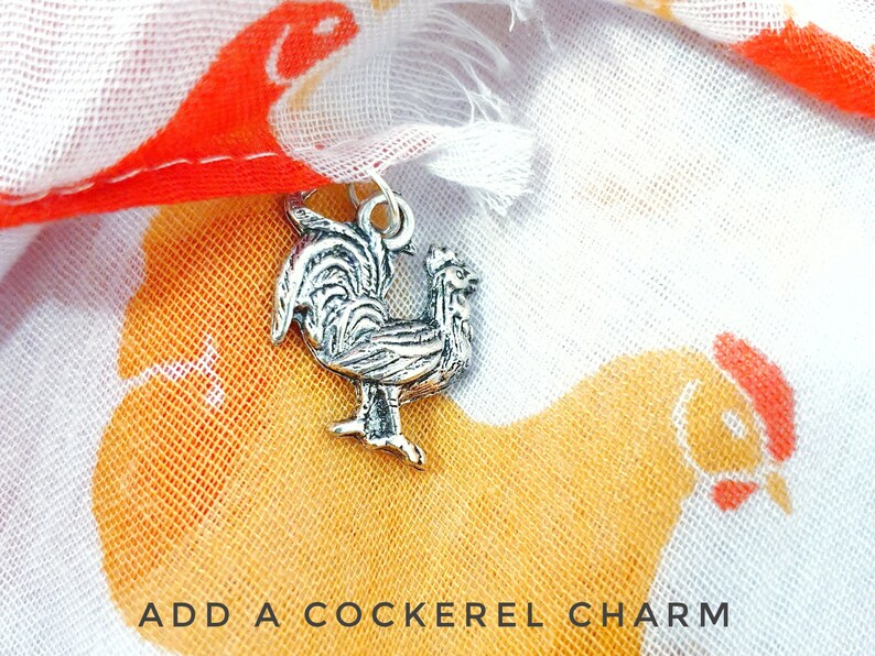 May include: A silver rooster charm with a detailed design, against a background of a white fabric with a chicken print. The text "ADD A COCKEREL CHARM" is visible at the bottom of the image.