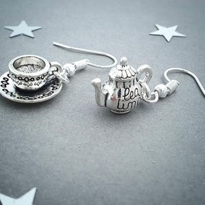 May include: Silver-toned earrings featuring a teacup and saucer charm paired with a teapot charm. The teapot charm has the words "tea time" written on it. The earrings are on a gray background with star-shaped decorations.
