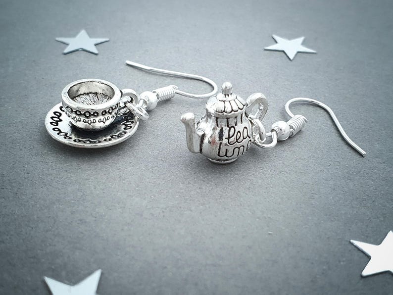 May include: Silver-toned earrings featuring a teacup and saucer charm paired with a teapot charm. The teapot charm has the words "tea time" written on it. The earrings are on a gray background with star-shaped decorations.