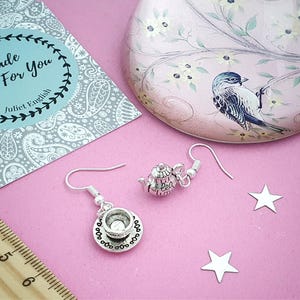 May include: Silver-toned teapot and teacup earrings with a matching design. The earrings are displayed on a pink surface with star-shaped confetti, a decorative tin, and a tag that reads "Made Just For You Juliet English."